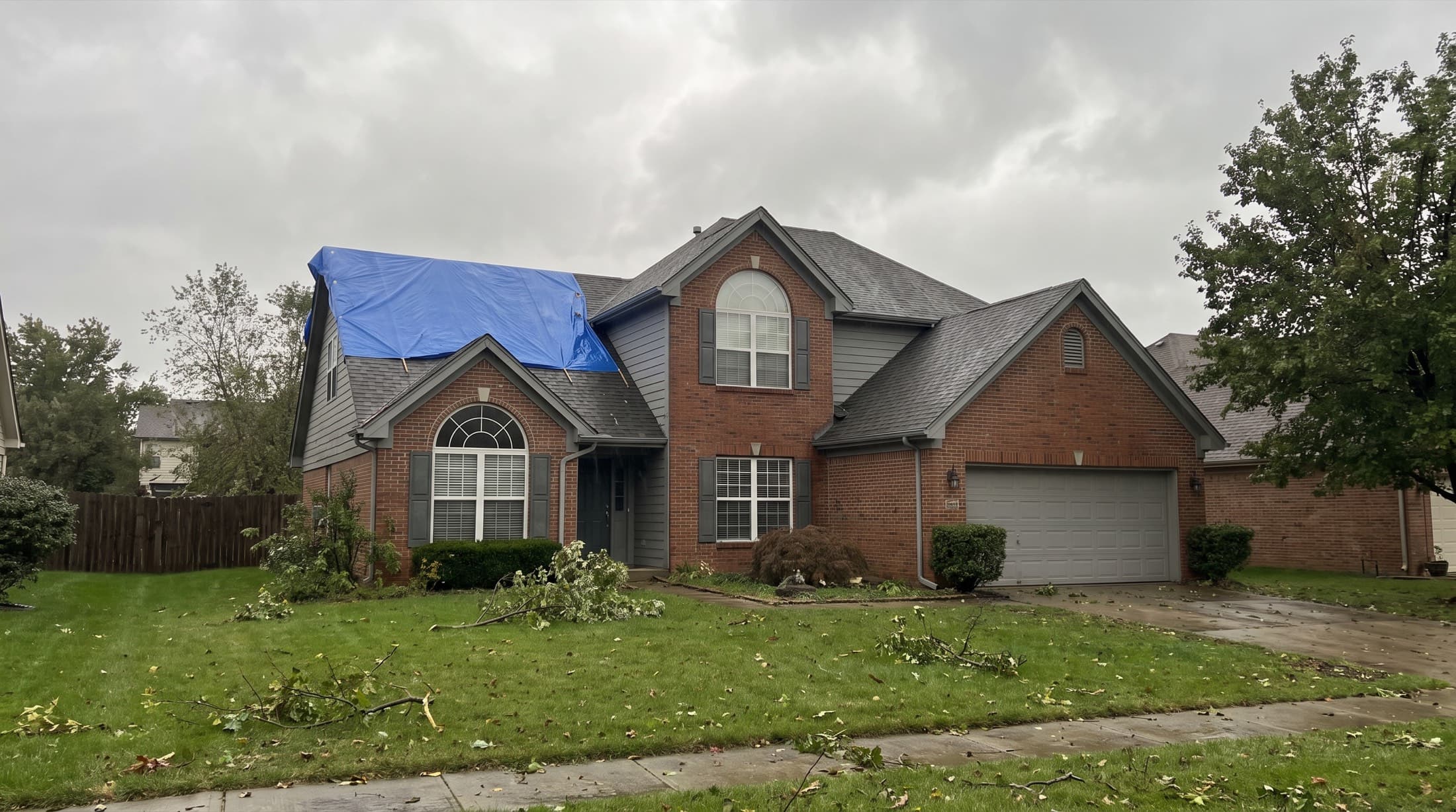 A two-story home the morning after a storm, an emergency tarp stretched over one roof slope and branches on the lawn
