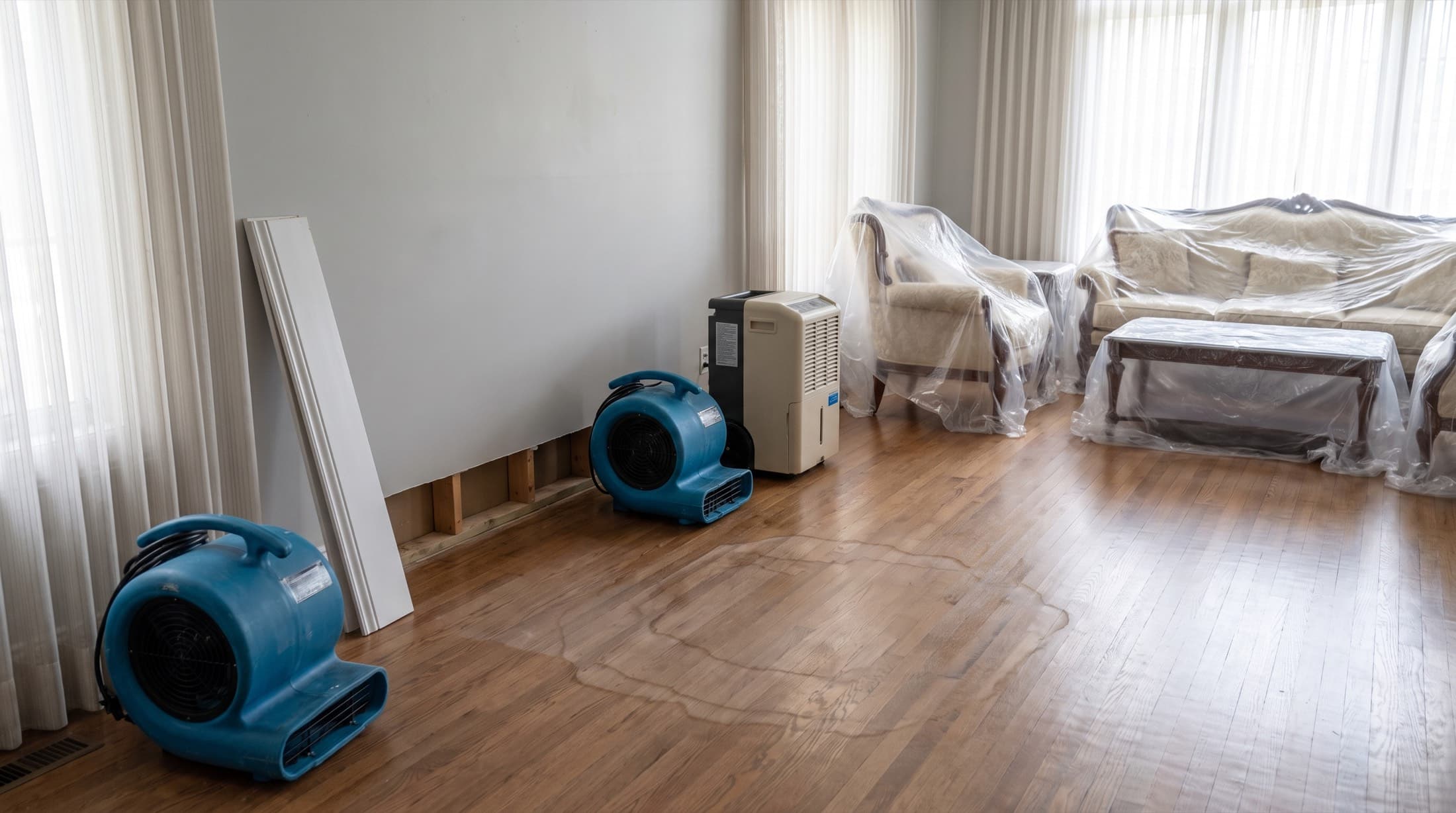 Air movers and a dehumidifier drying a living room, baseboard pulled and furniture protected under plastic